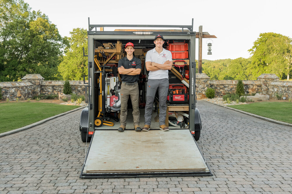 Kade Bloom's black and red KORE workshop trailier has the door open and laying as a ramp on the ground, displaying a shop full of tools, and he and his employee Nathaniel are standing together at the door smiling with arms crossed.